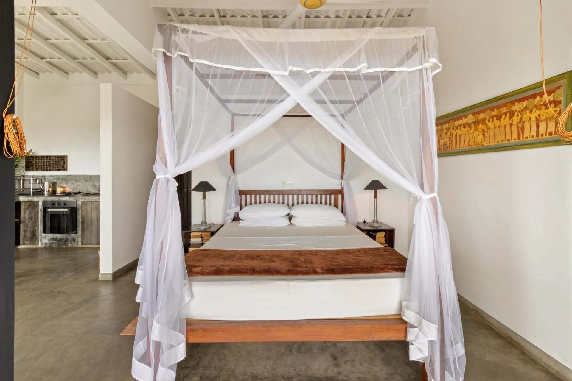 Four-poster bed with white mosquito net canopy, wooden frame, and brown throw blanket in spacious room with concrete floor, bedside lamps, and kitchen area in background.