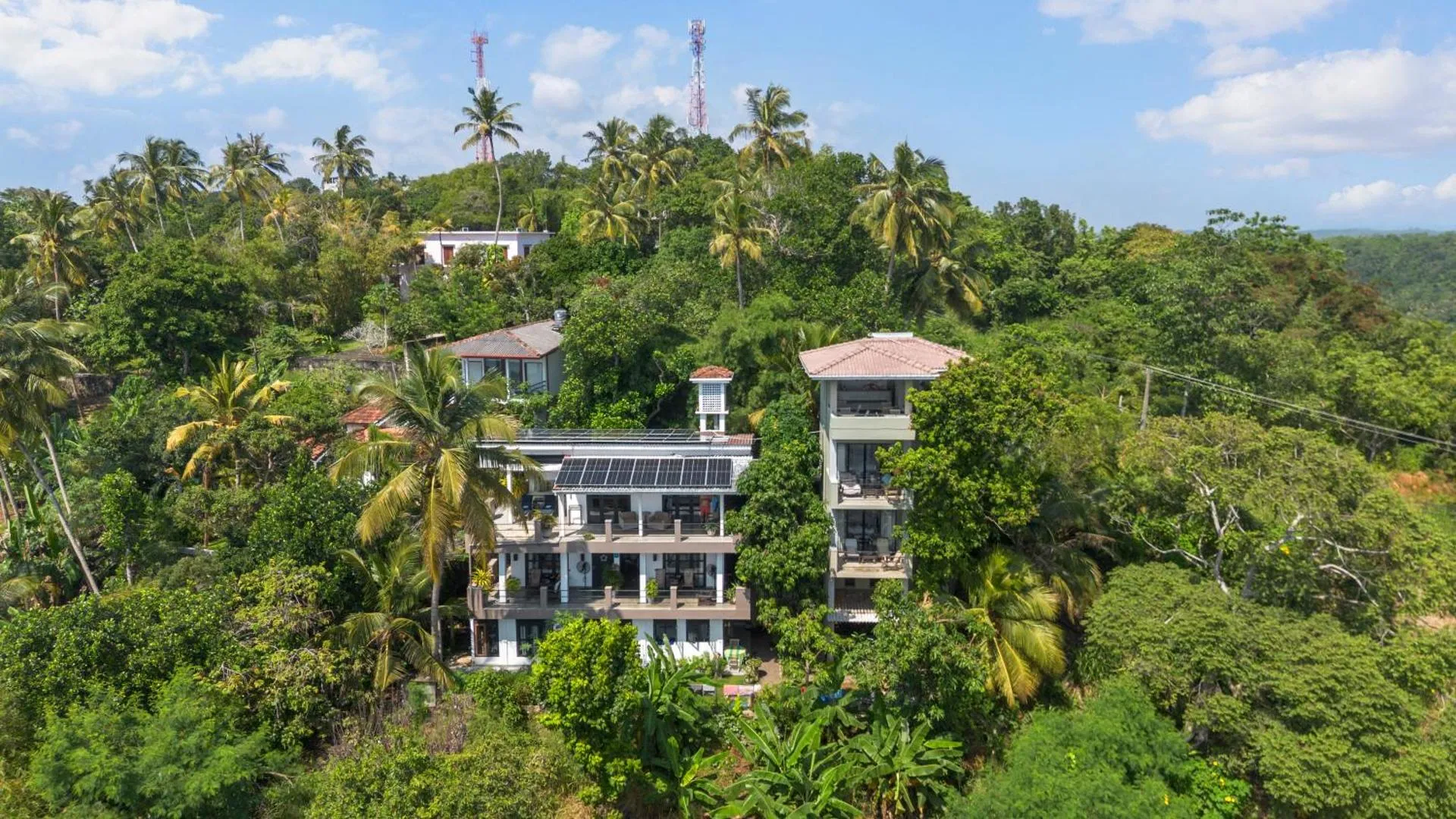 Aerial view of a multi-story building with solar panels nestled among lush greenery and palm trees on a hillside under a partly cloudy sky.