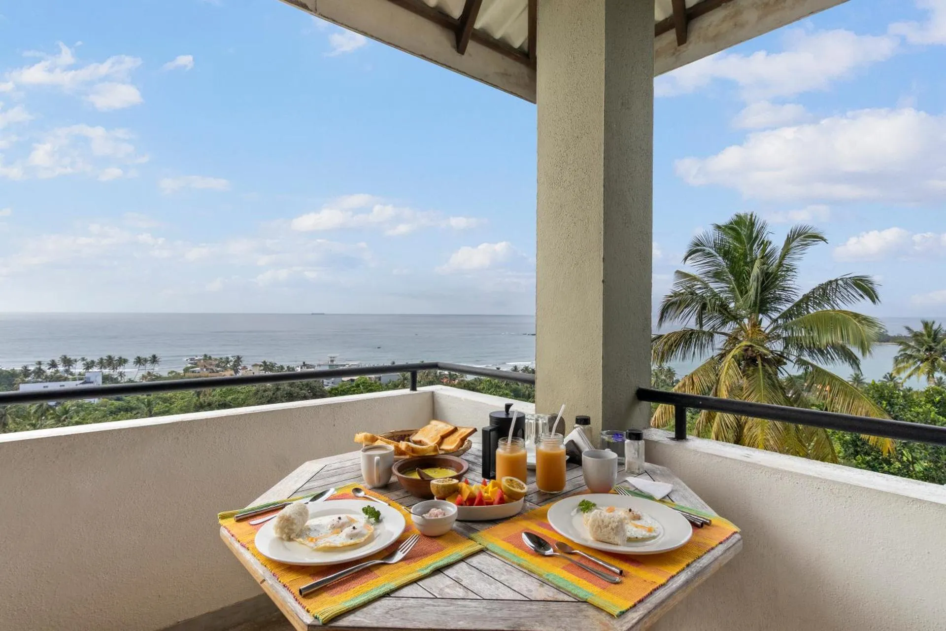 Breakfast spread on a wooden table on a balcony overlooking the ocean, with tropical palm trees and a clear blue sky in the background.