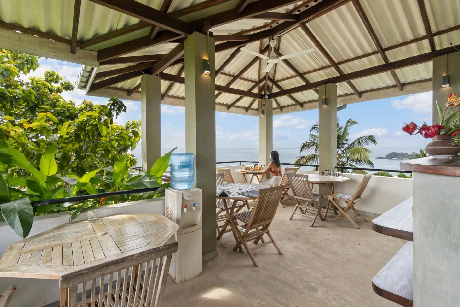 Outdoor dining area with wooden tables and chairs under a corrugated metal roof, overlooking the ocean with lush greenery and palm trees in the background.