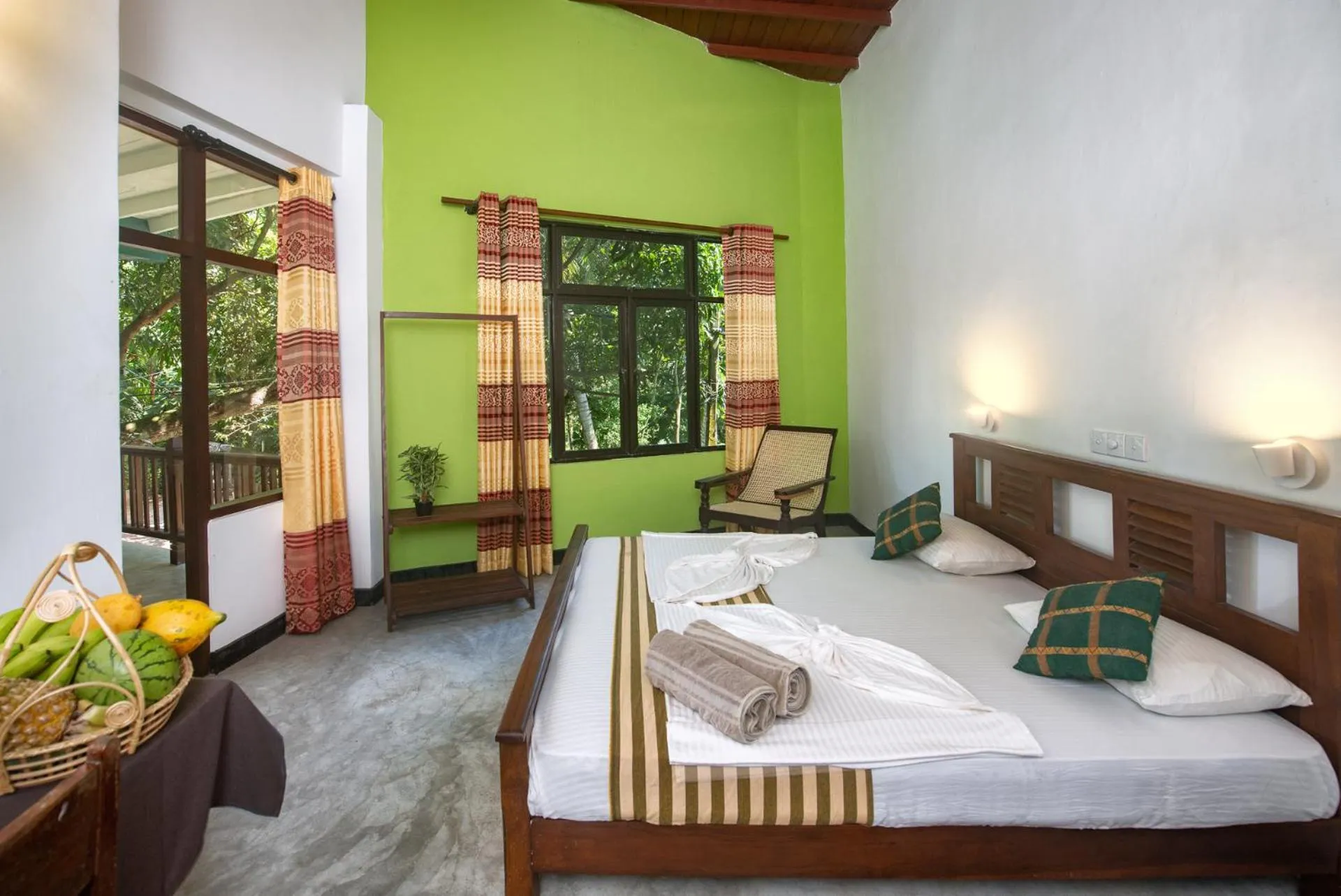 Bedroom with wooden bed, white linens, green accent wall, patterned curtains, wicker chair, fruit basket, and view to balcony with greenery.