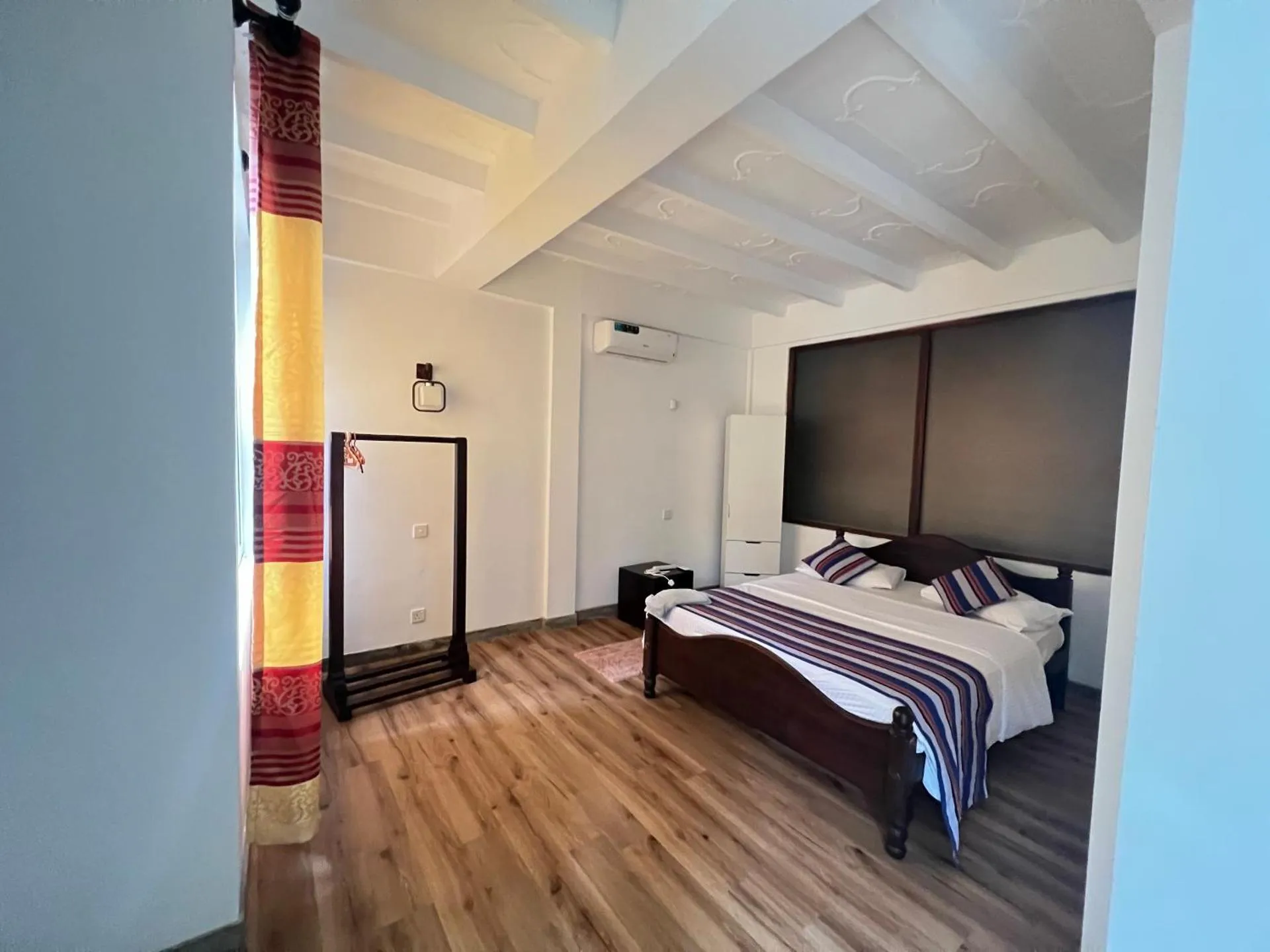 Cozy hotel room with wooden floor, white ceiling beams, single bed with striped bedding, full-length mirror, and colorful curtain near entrance.