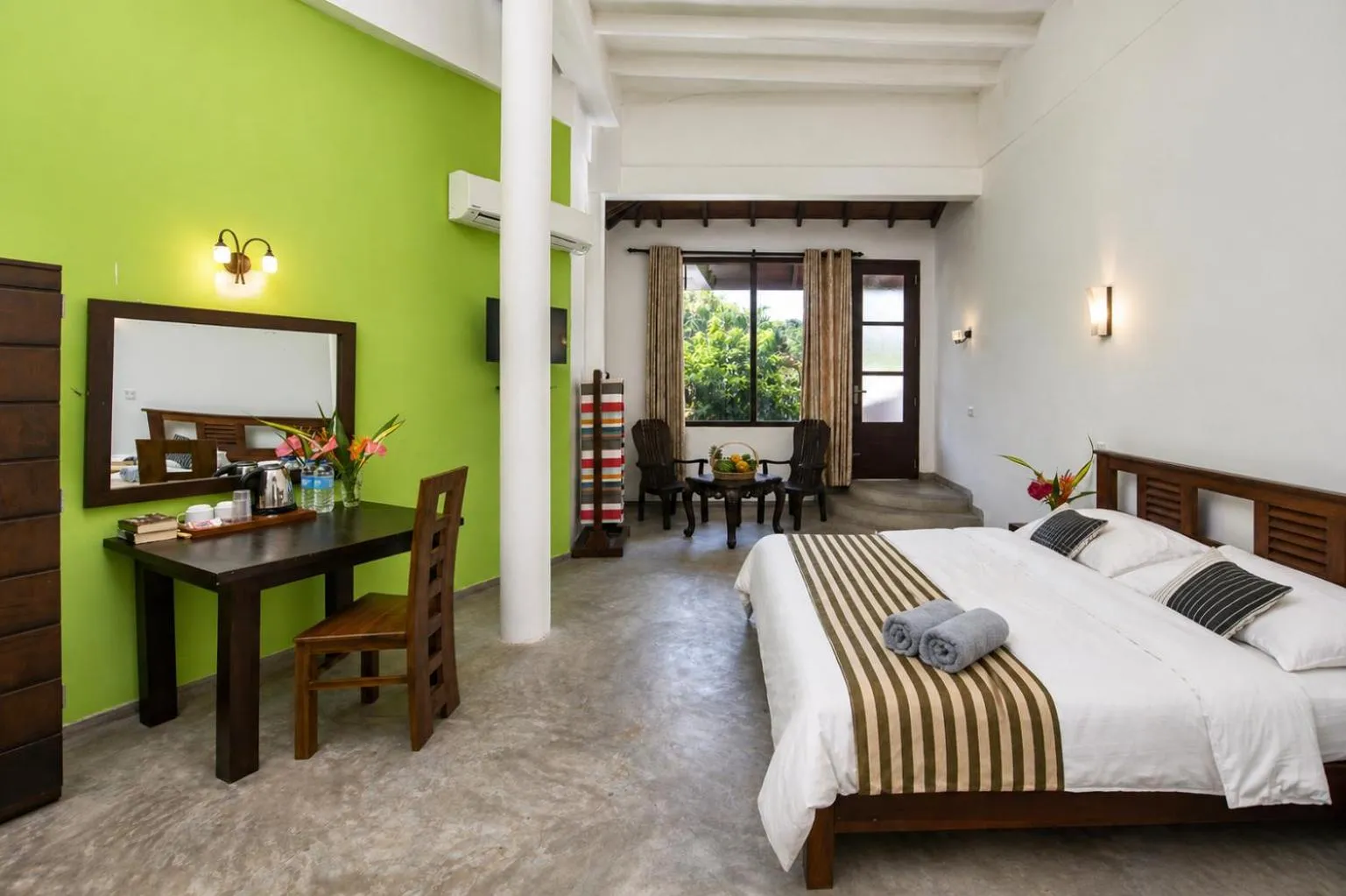 Hotel room with lime green accent wall, wooden bed with white linens and striped runner, dark wood desk and chair, mirror, and seating area near window with outdoor view.