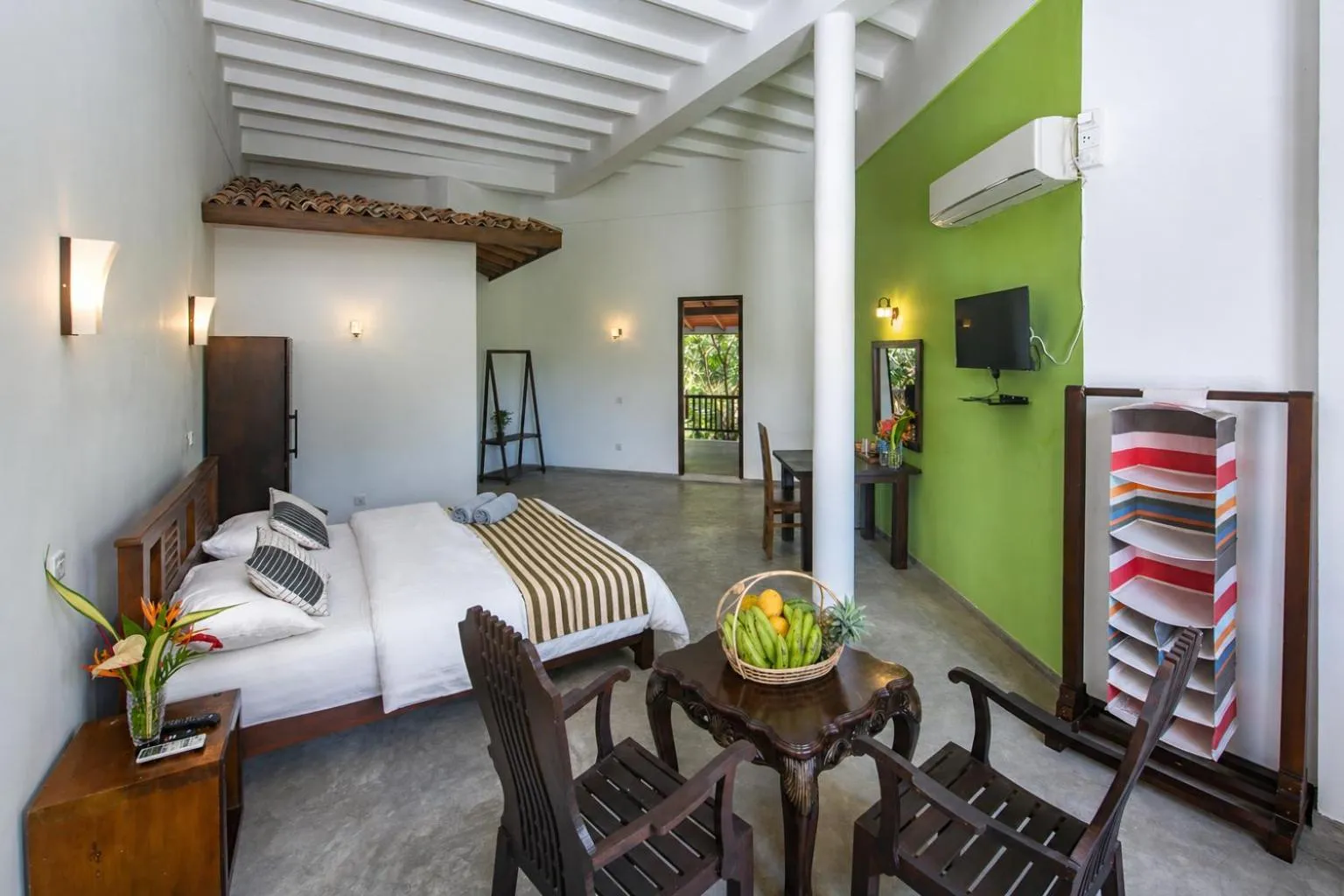 Bright hotel room with white bed, wooden furniture, green accent wall, ceiling fan, flat-screen TV, and fruit basket on table.