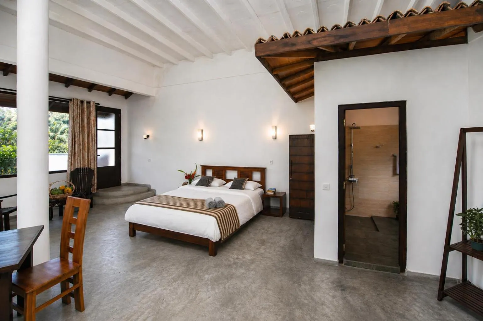 Spacious hotel room with white walls, wooden beams, and concrete floor, featuring a king-sized bed with striped throw, wooden furniture, and an open doorway to a walk-in shower.
